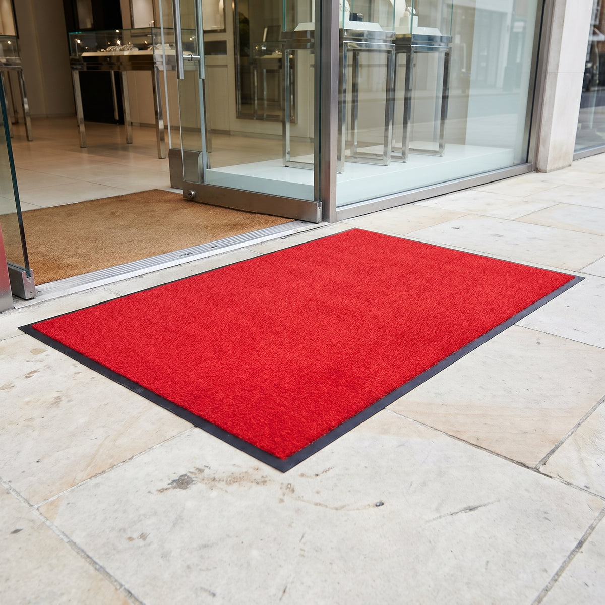 Red outdoor brush entrance mat with black rubber border outside a designer boutique