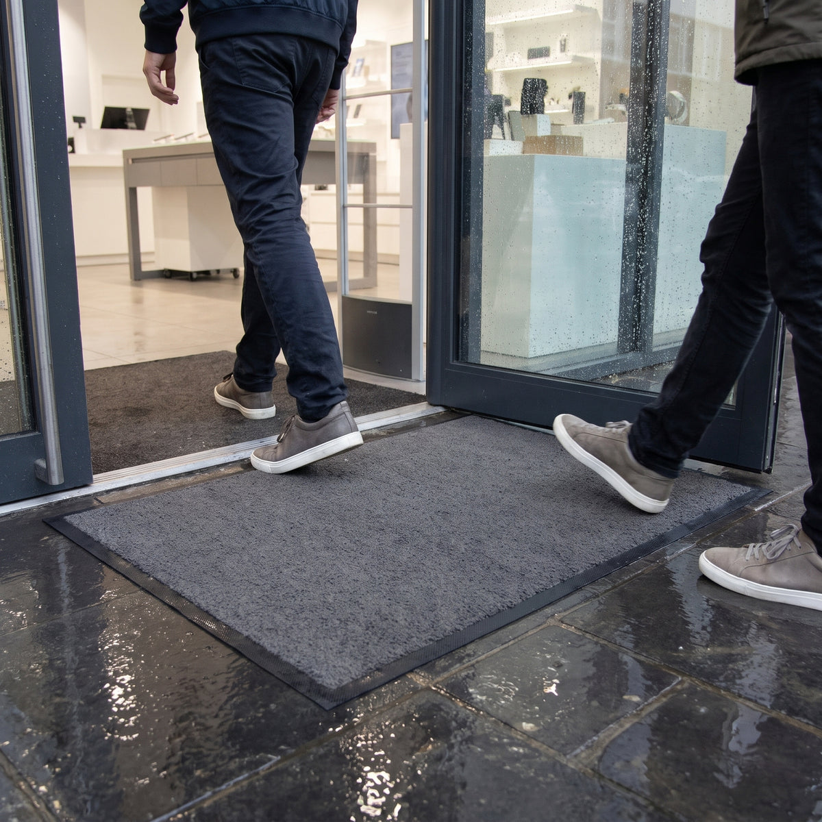 Customers stepping onto a grey outdoor brush mat at a shop doorway on a rainy day