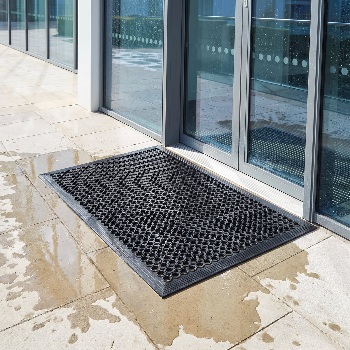 Outdoor rubber drainage mat staying clear on wet paving outside an office entrance after rain