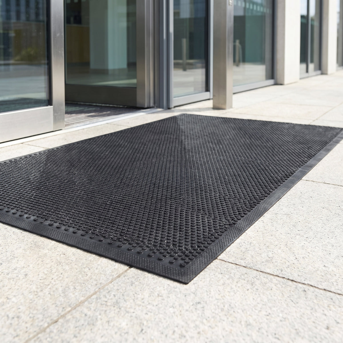 Black low-profile outdoor rubber scraper door mat outside a corporate glass entrance in daylight
